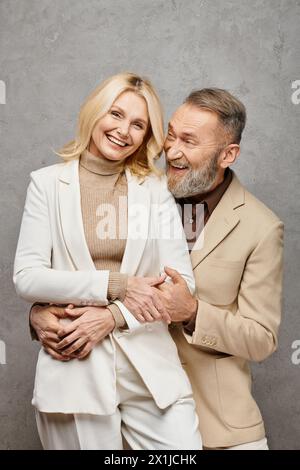 Un'elegante coppia matura con abiti in debonair che condivide un caldo e tenero abbraccio su uno sfondo grigio. Foto Stock
