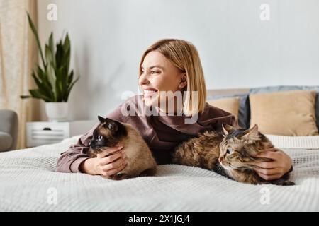 Una donna con i capelli corti si rilassa su un letto con due gatti al suo fianco, godendosi un momento tranquillo a casa. Foto Stock