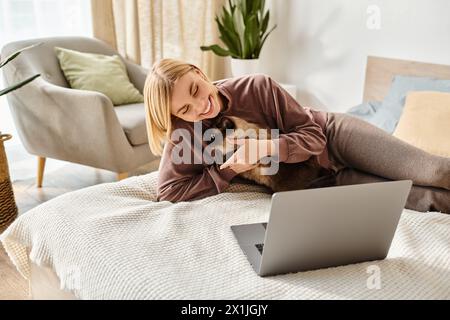 Una donna elegante con capelli corti si rilassa su un letto, avvolto nel suo portatile mentre il suo gatto si coccola accanto a lei. Foto Stock