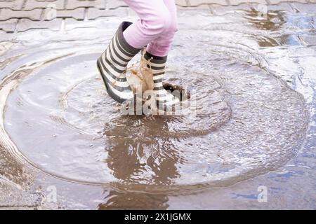 primo piano, la bambina di 5 anni salta gioiosamente in un pozzetto indossando stivali di gomma, i piedi dei bambini in schizzi d'acqua, saltando felicemente, i piaceri dell'infanzia Foto Stock