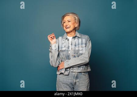 In uno studio, una donna matura con i capelli grigi si erge con sicurezza su uno sfondo blu. Indossa un outfit alla moda in denim e irradia felicità Foto Stock