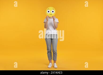 Donna che copre il viso con emoticon sorpresa su sfondo giallo Foto Stock