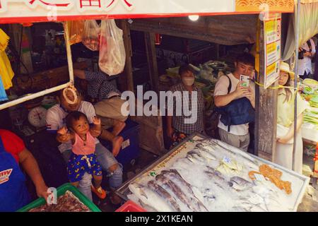 Maeklong, Thailandia - Vista dalla carrozza mentre l'uomo usa lo smartphone per catturare il treno passando il pescivendolo nel mercato dei treni molto vicino alle bancarelle Foto Stock