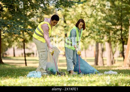 Una coppia socialmente attiva e diversificata di giubbotti di sicurezza e guanti si erge unita nell'erba lussureggiante, pulendo con passione il parco. Foto Stock