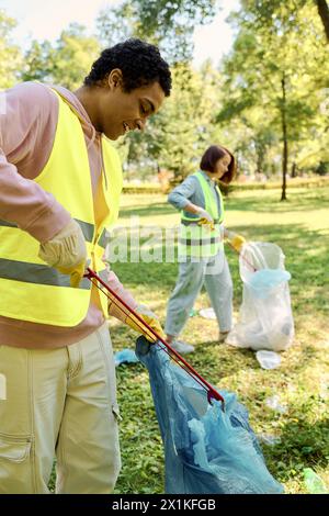 Socialmente attiva, coppia diversificata di giubbotti di sicurezza e guanti che puliscono il parco insieme a sacchi per la spazzatura. Foto Stock