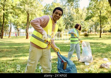 Un uomo afroamericano in un giubbotto di sicurezza giallo tiene una borsa blu mentre pulisce in un parco con sua moglie Foto Stock