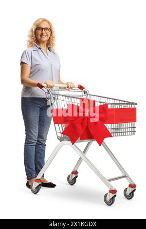 Donna matura in piedi con un carrello della spesa vuoto decorato con un fiocco rosso isolato su sfondo bianco Foto Stock