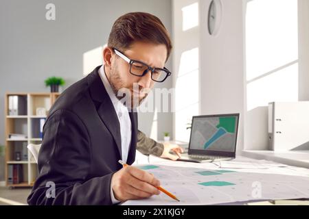 Ispettore catastale seduto in ufficio, che lavora con le mappe catastali e utilizza un computer portatile Foto Stock