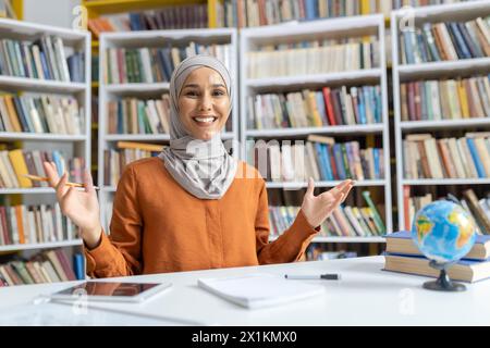 Fiduciosa e allegra maestra musulmana in un hijab che sorride alla telecamera mentre si trova in una biblioteca con scaffali pieni di libri. Foto Stock