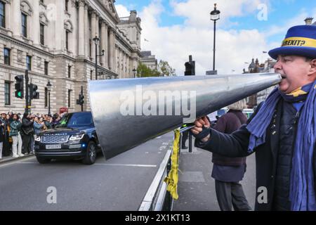 Londra, Regno Unito. 17 aprile 2024. Steve Bray, meglio conosciuto come "Stop Brexit Man" di Westminster, grida all'auto del PM con il suo megafono proprio mentre il corteo di Rishi Sunak li passa davanti per il PM per partecipare ai PMQ al Parlamento. A un certo punto, il megafono sembra "ingoiare" l'auto del PM in una divertente illusione ottica. Crediti: Imageplotter/Alamy Live News Foto Stock