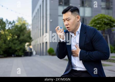 Un giovane asiatico che soffre di tosse e asma in una tuta da lavoro è seduto vicino a un edificio di uffici, tenendo le mani al petto e usando un inalatore. Foto Stock