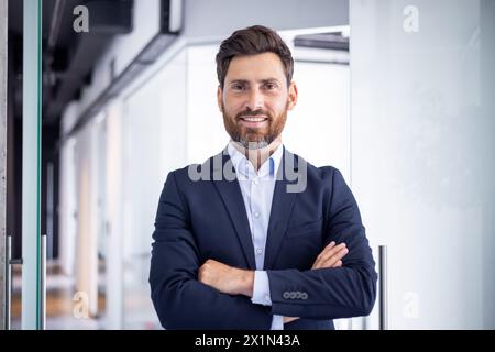 Ritratto di un giovane uomo d'affari sorridente e di successo in piedi in ufficio, che incrocia le braccia sul petto e guarda la fotocamera. Foto Stock