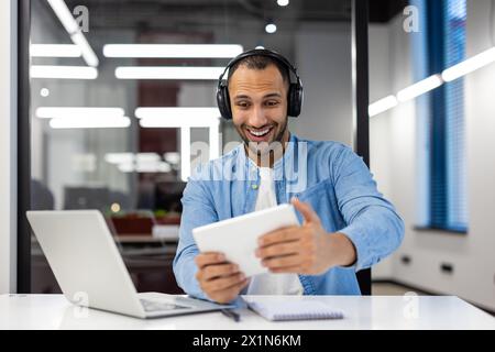 Sorridente giovane latinoamericano seduto a una scrivania in ufficio con le cuffie e divertiti a giocare online su un tablet. Foto Stock