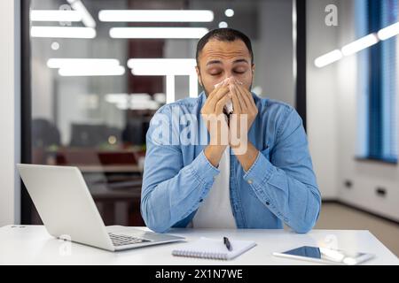 Un giovane latinoamericano lavora in ufficio, si siede a una scrivania e soffre di febbre virale e allergie, si pulisce il naso con un tovagliolo da un naso che cola. Foto Stock