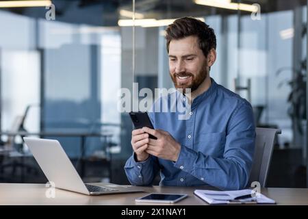 Un giovane lavoratore in ufficio sorridente si siede in ufficio a una scrivania e utilizza un telefono cellulare, scrive un messaggio, corrisponde. Foto Stock