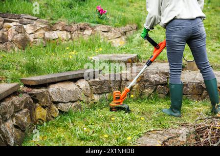La donna sta rifilando l'erba accanto al muro di pietra in giardino. Cura del prato con tagliaerba Foto Stock