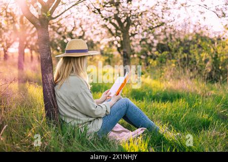 Donna rilassata seduta sotto un albero di ciliegio in fiore e leggendo un libro per migliorare la sua consapevolezza. Godetevi momenti di solitudine e relax in primavera Foto Stock