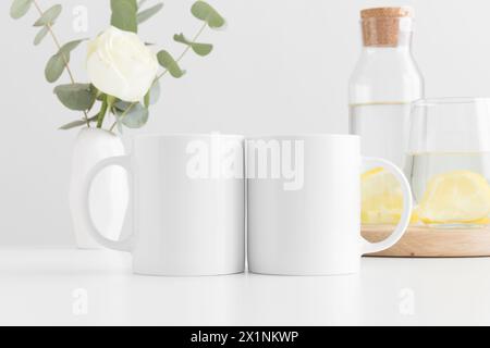 Due tazze con fiori in un vaso, un bicchiere e una bottiglia su un tavolo bianco. Foto Stock