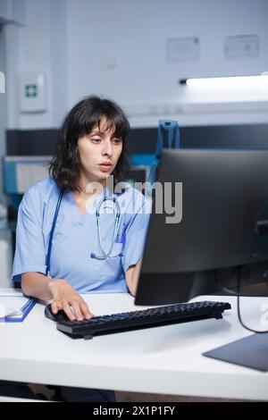 Al banco dell'ufficio della clinica, un'infermiera caucasica determinata controlla e analizza le sue note. Primo piano di medico in scrub blu preparandosi a vedere le persone per le visite mediche. Foto Stock