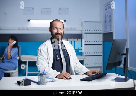 Medico di mezza età sorridente alla sua scrivania in uno studio medico. Il medico caucasico maschile sembra indossare un camice da laboratorio bianco, l'infermiera è sullo sfondo durante una chiamata. Scatto verticale. Foto Stock
