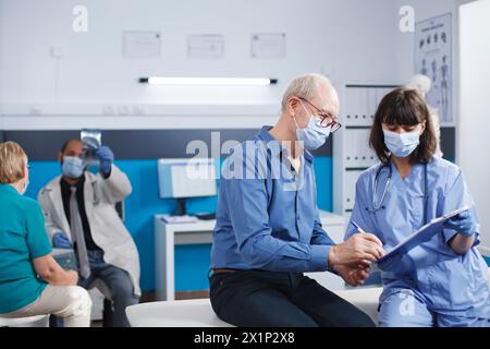 Medico specialista con appunti e documento ospedaliero per il paziente di sesso maschile anziano da firmare. L'infermiera e l'anziano si sedevano sul letto della clinica, entrambi indossavano maschere facciali, guardando i documenti dell'assicurazione clinica. Foto Stock
