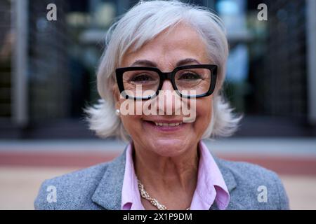 Ritratto di donna d'affari caucasica matura con occhiali e capelli bianchi in tuta formale sorridente Foto Stock
