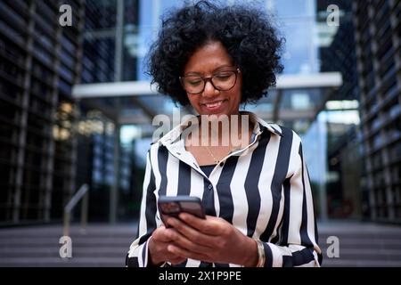 Donna afroamericana adulta in tuta formale in piedi di fronte all'edificio degli uffici utilizzando il telefono cellulare Foto Stock