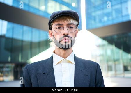 Un giovane hipster caucasico con un'elegante tuta blu, cappello e occhiali in piedi seri e sicuri di sé Foto Stock