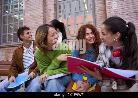 Gruppo sorridente di giovani studenti seduti sulla panchina che studiano insieme fuori dall'edificio universitario Foto Stock