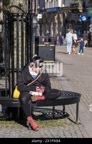 17 aprile 2024. Elgin, Moray, Scozia. Si tratta di una giovane signora seduta su una panchina all'interno dell'area pedonale di High Street a Elgin. Sta ascoltando Foto Stock