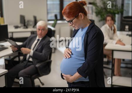 Una donna incinta soffre di dolore e tiene lo stomaco in piedi nel mezzo dell'ufficio accanto ai suoi colleghi. Foto Stock