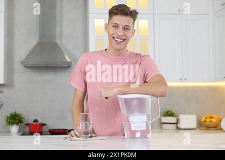 Uomo felice con caraffa filtro e bicchiere d'acqua pulita a tavola in cucina Foto Stock