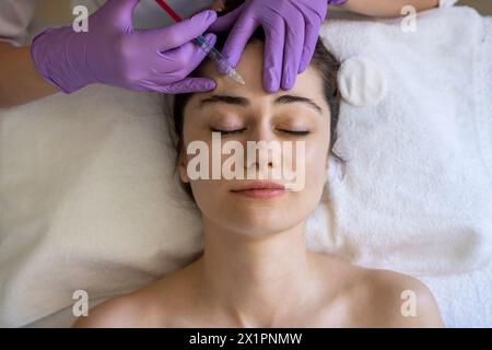 Vista dall'alto di una donna che riceve un'iniezione di riempitivo botox anti-invecchiamento sulla fronte da un cosmetologo professionista nel salone di bellezza. Trattamento viso e. Foto Stock