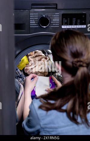Donna seduta di fronte a una lavatrice che carica abiti sporchi, tovaglioli per lavare abiti colorati Foto Stock