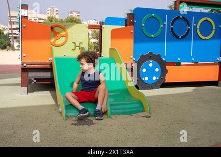 Ragazzo solitario con capelli ricci seduto su uno scivolo colorato. Foto Stock