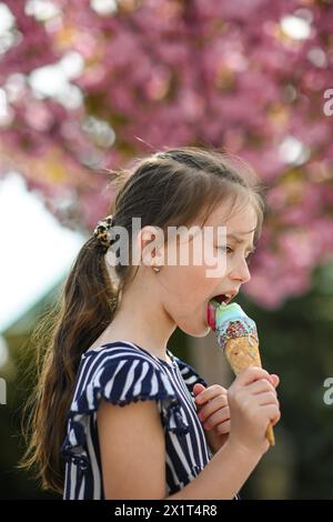 La bambina lecca il gelato in estate. Bellissimo albero rosa in fiore sullo sfondo-bokeh. Foto Stock