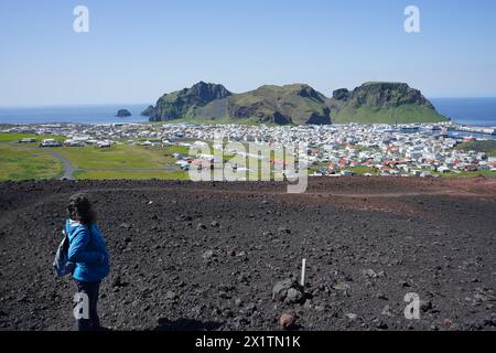 Una giovane donna guarda la città di Heimaey e la montagna di Heimaklettur dalla cima del cono vulcanico di Eldfell, le isole Westman, Islanda Foto Stock