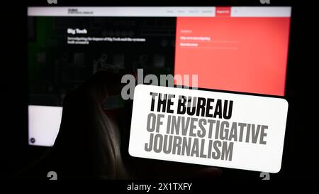 Persona con smartphone e logo dell'organizzazione il Bureau of Investigative Journalism (TBIJ) davanti al sito web. Mettere a fuoco il display del telefono. Foto Stock