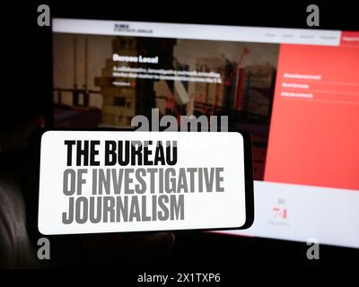 Persona che detiene un telefono cellulare con logo dell'organizzazione il Bureau of Investigative Journalism (TBIJ) di fronte alla pagina web. Mettere a fuoco il display del telefono. Foto Stock