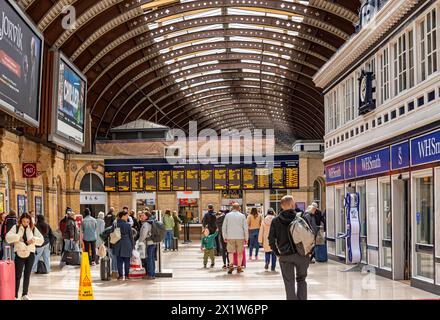 Un baldacchino di ferro copre un atrio della stazione ferroviaria con un cartello elettronico che mostra le informazioni sui treni. Un negozio è su un lato e una folla di persone Foto Stock