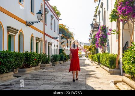 Una donna con un vestito rosso cammina lungo una strada stretta con un cappello. La strada è costeggiata da alberi e case, e ci sono piante in vaso lungo il lato Foto Stock