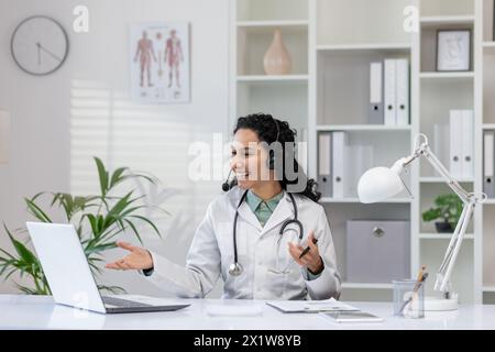 Un medico femminile professionista con uno stetoscopio impegnato in una consultazione online interattiva, sorridendo calorosamente in uno studio medico ben attrezzato. Foto Stock