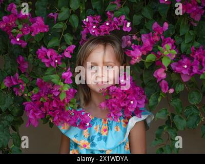Adorabile bambina in abito bianco ridendo e guardando via mentre si trova in piedi tra bellissimi fiori in un prato incredibile Foto Stock