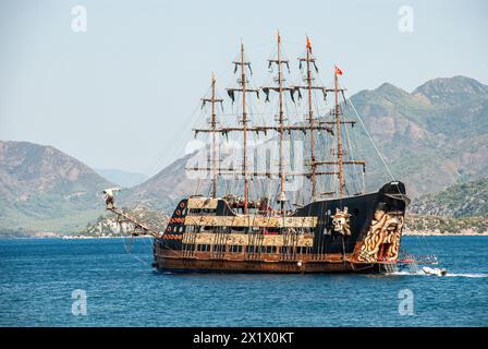 Una divertente nave pirata con le sue vele nere abbassate, che trasportano i turisti che si divertono a bordo, naviga nel tranquillo porto circondato da verdi colline Foto Stock