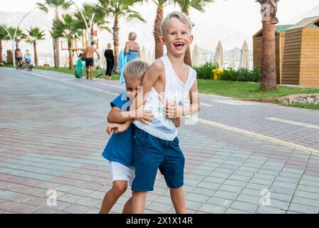 Due giovani ragazzi ridono e giocano a una partita di lotta sul lungomare lastricato, con palme sullo sfondo e pedoni che camminano Foto Stock