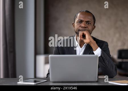 Uomo adulto attento in abiti rigidi seduti accanto a un computer portatile e sfregando il mento con il braccio in casa. Persona afroamericana che guarda la telecamera e si sente confusa con la soluzione del problema attuale. Foto Stock