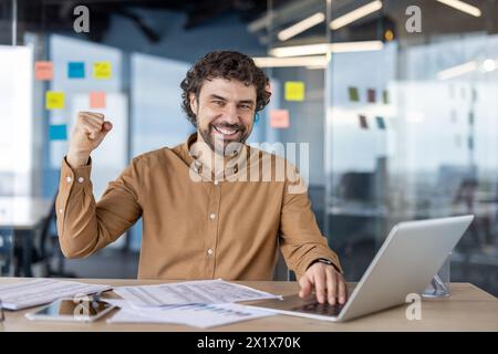 Allegro freelance che tiene la mano sulla tastiera del laptop sorridendo alla fotocamera e alzando la mano con il pugno serrato. Un creatore di successo si accontenta di concludere un accordo con un cliente a lungo termine. Foto Stock