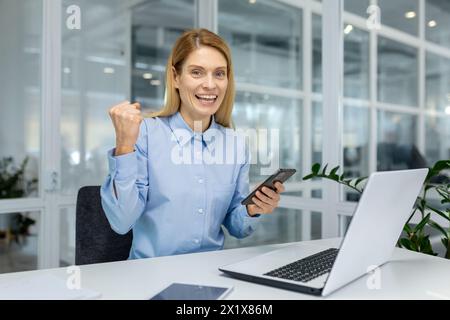 Donna in camicia positiva che fa un gesto trionfale con la mano tenendo lo smartphone in un ambiente di lavoro moderno. Un manager allegro che riceve un piacevole messaggio di testo con buone notizie sulle opportunità di lavoro. Foto Stock