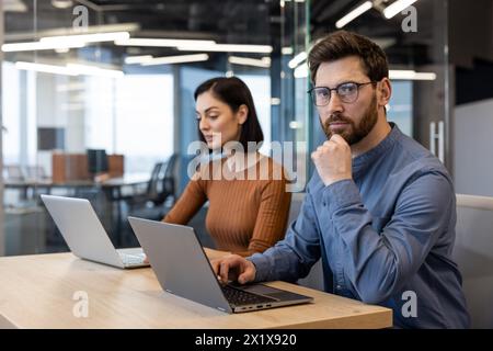 Pensivo ragazzo non rasato in camicia di denim che poggia la testa sulla mano mentre guarda la fotocamera accanto a una collega. Due designer digitali sviluppano una nuova strategia creativa per i social media per il team di marketing. Foto Stock