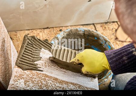 La Malta cementizia dell'adesivo per piastrelle viene applicata alle piastrelle utilizzando una cazzuola intagliata. Foto Stock